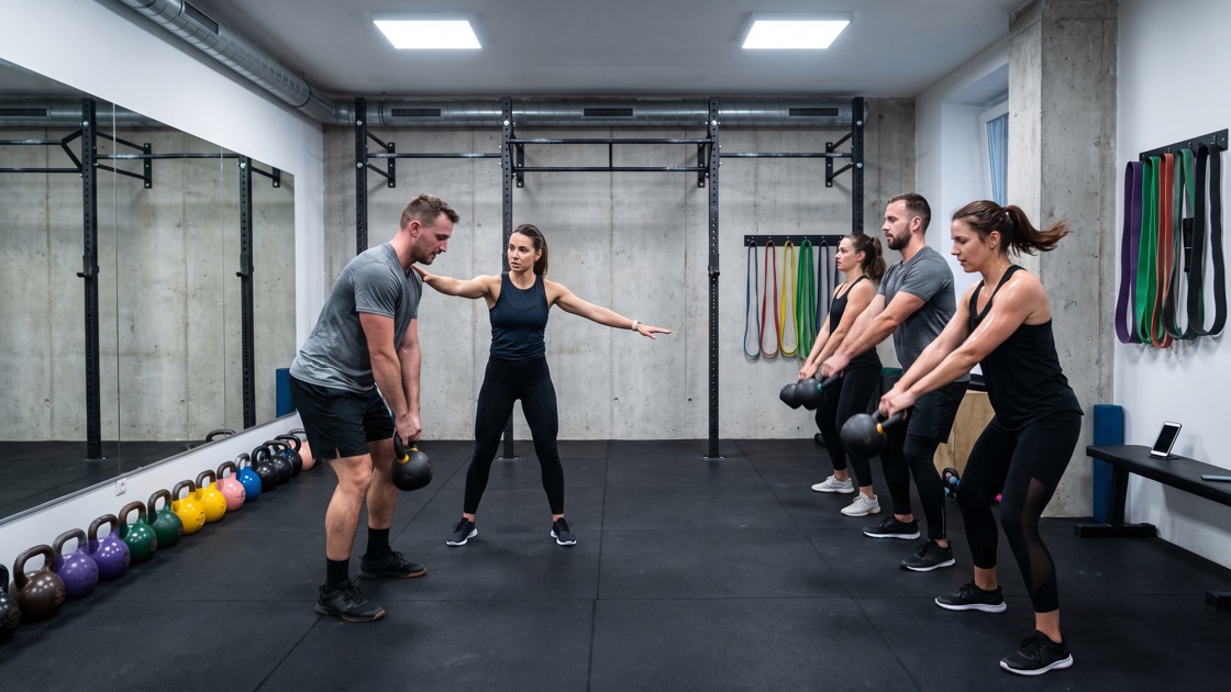 Edző javítja egy résztvevő tartását kettlebell-óra közben egy betonfalú stúdióban